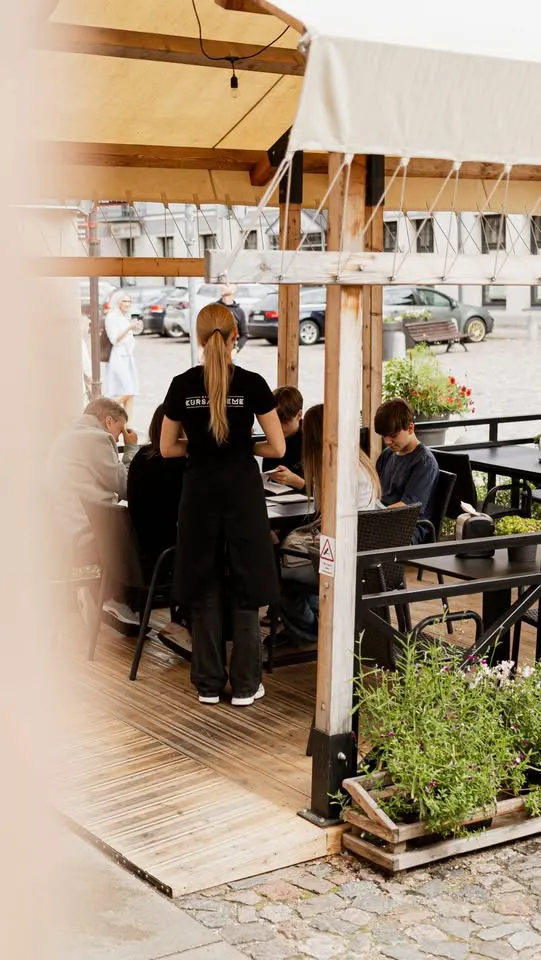 Terase ar koka nojumi un puķu kastēm, viesi pie galdiņiem