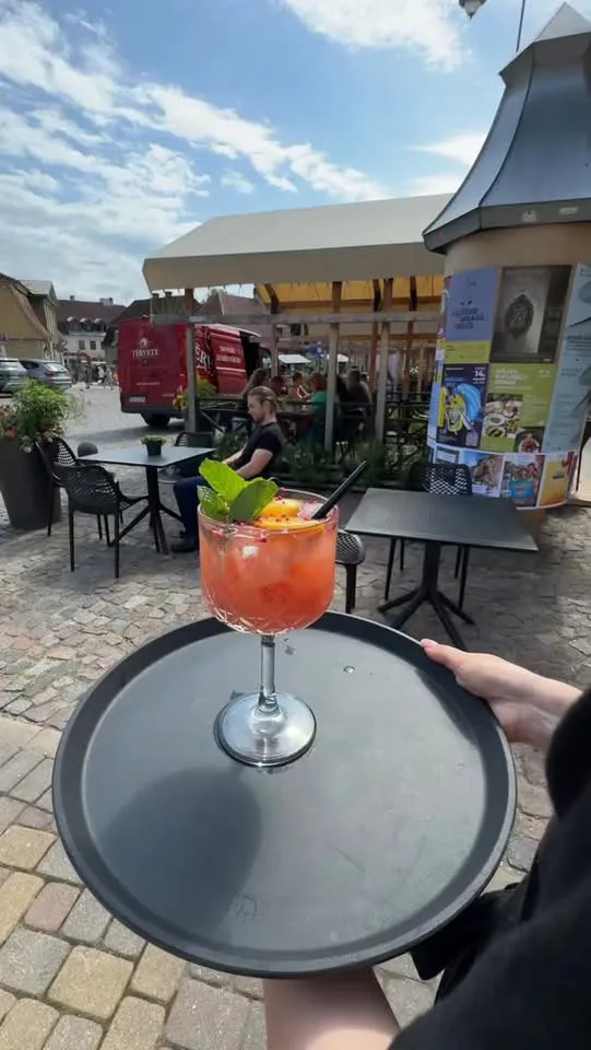 Oranžs kokteilis uz paplātes, fonā terase un vecpilsētas bruģis
